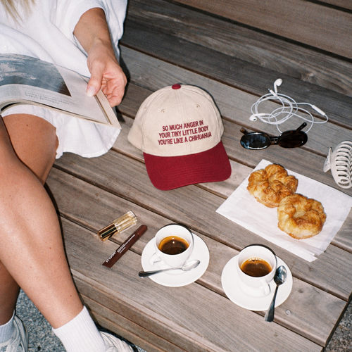 A lifestyle flat-lay on a wooden bench featuring a beige and red baseball cap with the text 'SO MUCH ANGER IN YOUR TINY LITTLE BODY YOU'RE LIKE A CHICKANNA'. The scene is styled with two cups of coffee, croissants, sunglasses, earphones, and a person's legs reading a book.