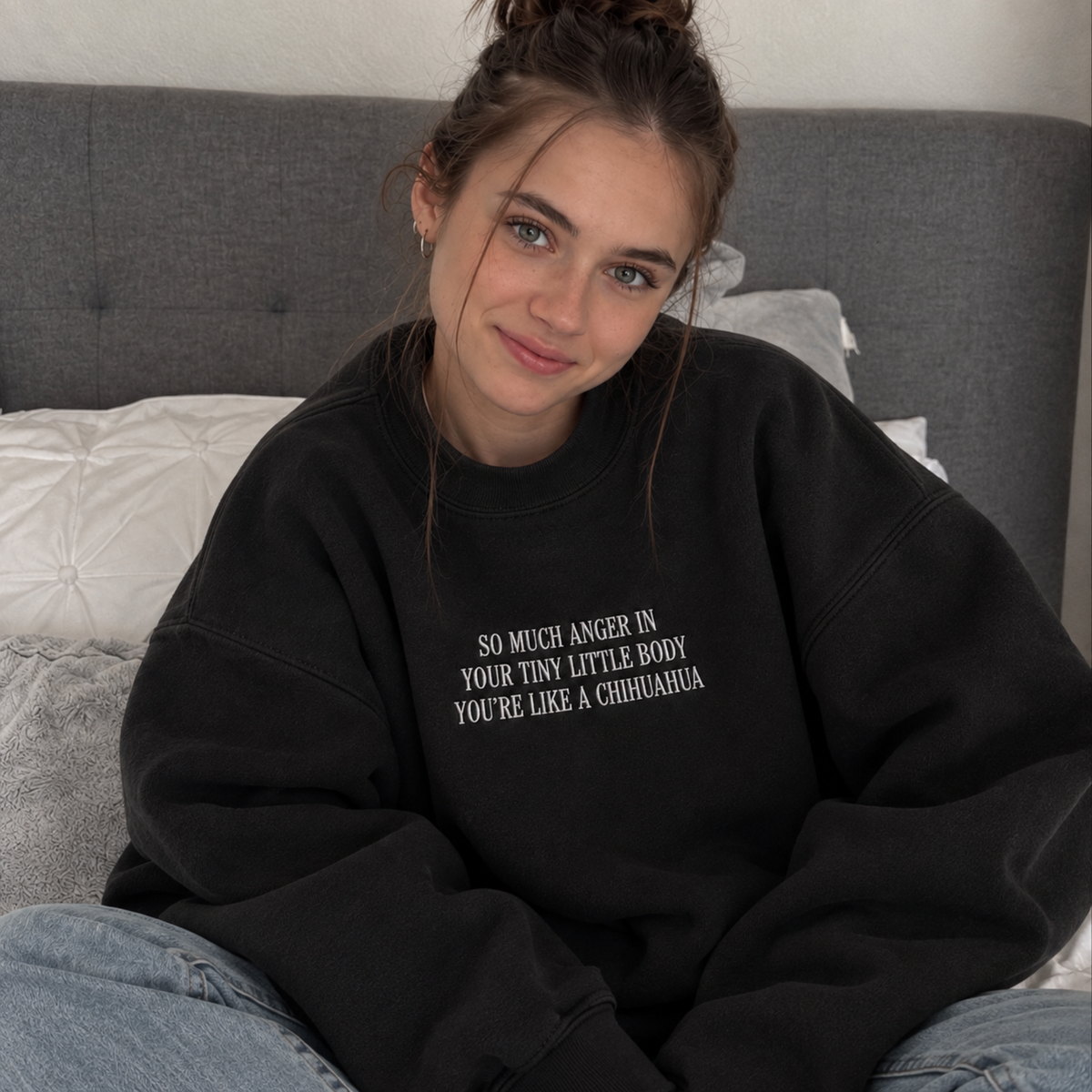 A young woman with her hair in a messy bun sits on a bed wearing a black oversized sweatshirt with white text that reads 'SO MUCH ANGER IN YOUR TINY LITTLE BODY YOU'RE LIKE A CHIHUAUA'.