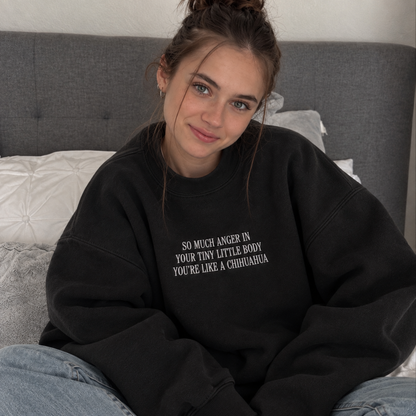 A young woman with her hair in a messy bun sits on a bed wearing a black oversized sweatshirt with white text that reads 'SO MUCH ANGER IN YOUR TINY LITTLE BODY YOU'RE LIKE A CHIHUAUA'.