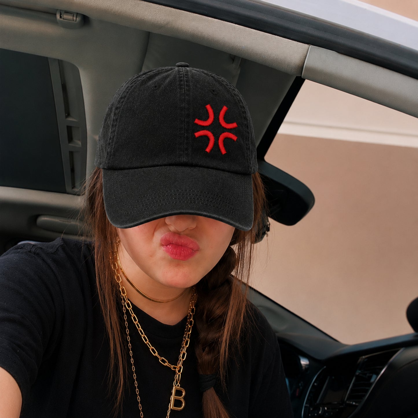 A person sitting inside a car wearing a black baseball cap with a red embroidered symbol on the front, a black t-shirt, and gold necklaces.
