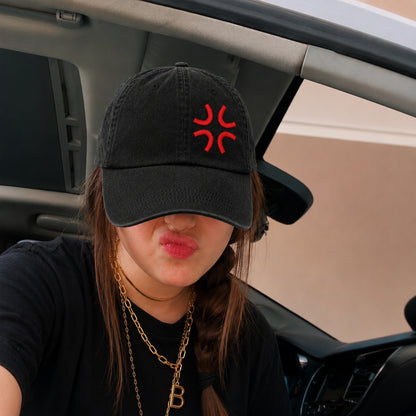 A person sitting inside a car wearing a black baseball cap with a red embroidered symbol on the front, a black t-shirt, and gold necklaces.