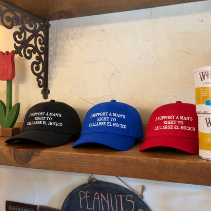 Three baseball caps in black, blue, and red are displayed on a wooden shelf. Each cap has white embroidered text that reads, 'I SUPPORT A MAN'S RIGHT TO CALLARSE EL HOCICO.