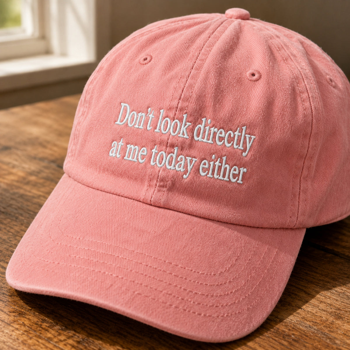 A dusty pink baseball cap with the embroidered text 'Don't look directly at me today either' in white, resting on a wooden table near a window.