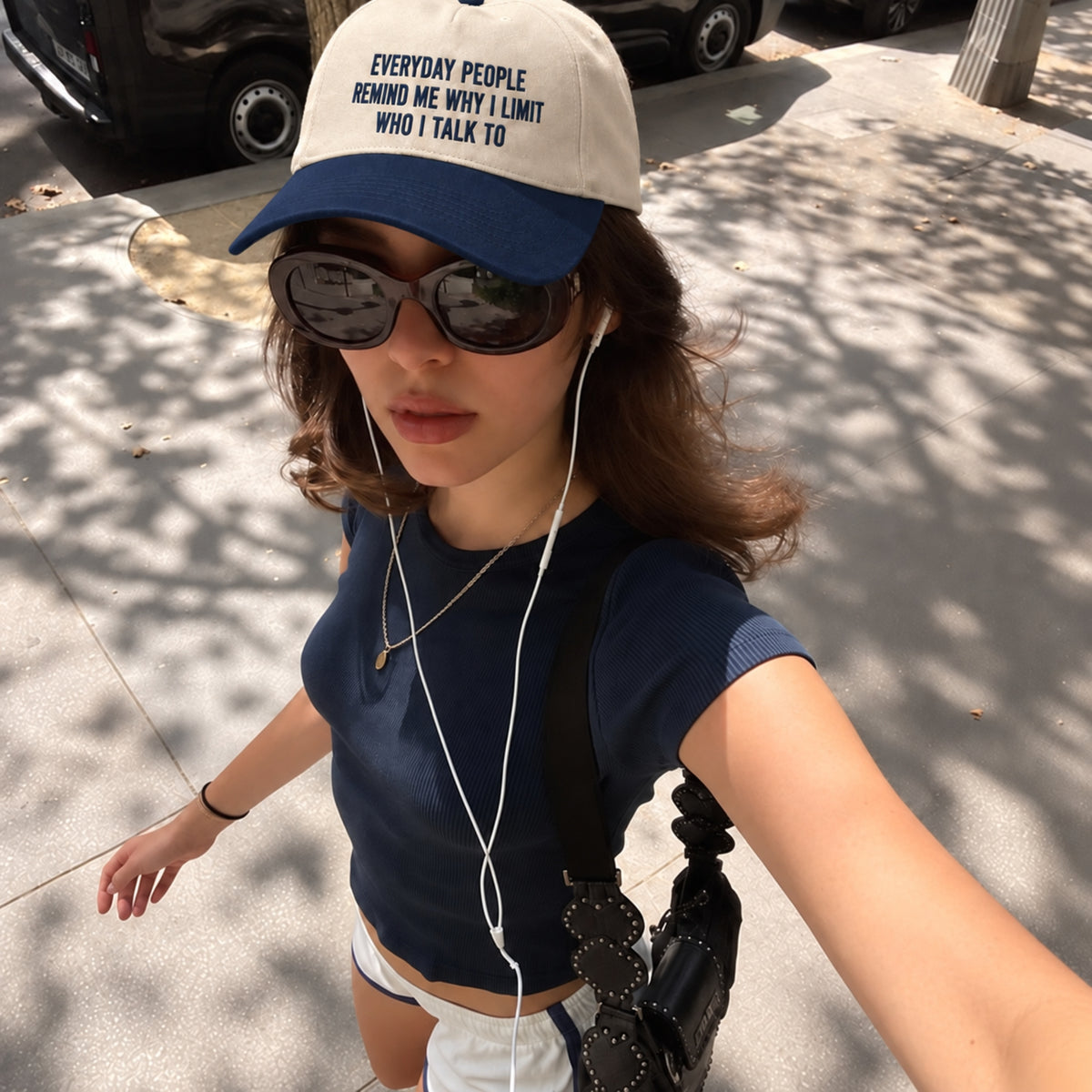 A woman takes a selfie outdoors wearing a navy blue and off-white baseball cap with the text 'EVERYDAY PEOPLE REMIND ME WHY I LIMIT WHO I TALK TO' in navy lettering. She is also wearing dark sunglasses, a navy ribbed short-sleeve crop top, white shorts, and carrying a black shoulder bag.