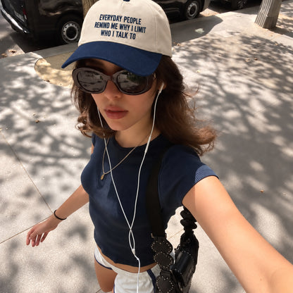 A woman takes a selfie outdoors wearing a navy blue and off-white baseball cap with the text 'EVERYDAY PEOPLE REMIND ME WHY I LIMIT WHO I TALK TO' in navy lettering. She is also wearing dark sunglasses, a navy ribbed short-sleeve crop top, white shorts, and carrying a black shoulder bag.