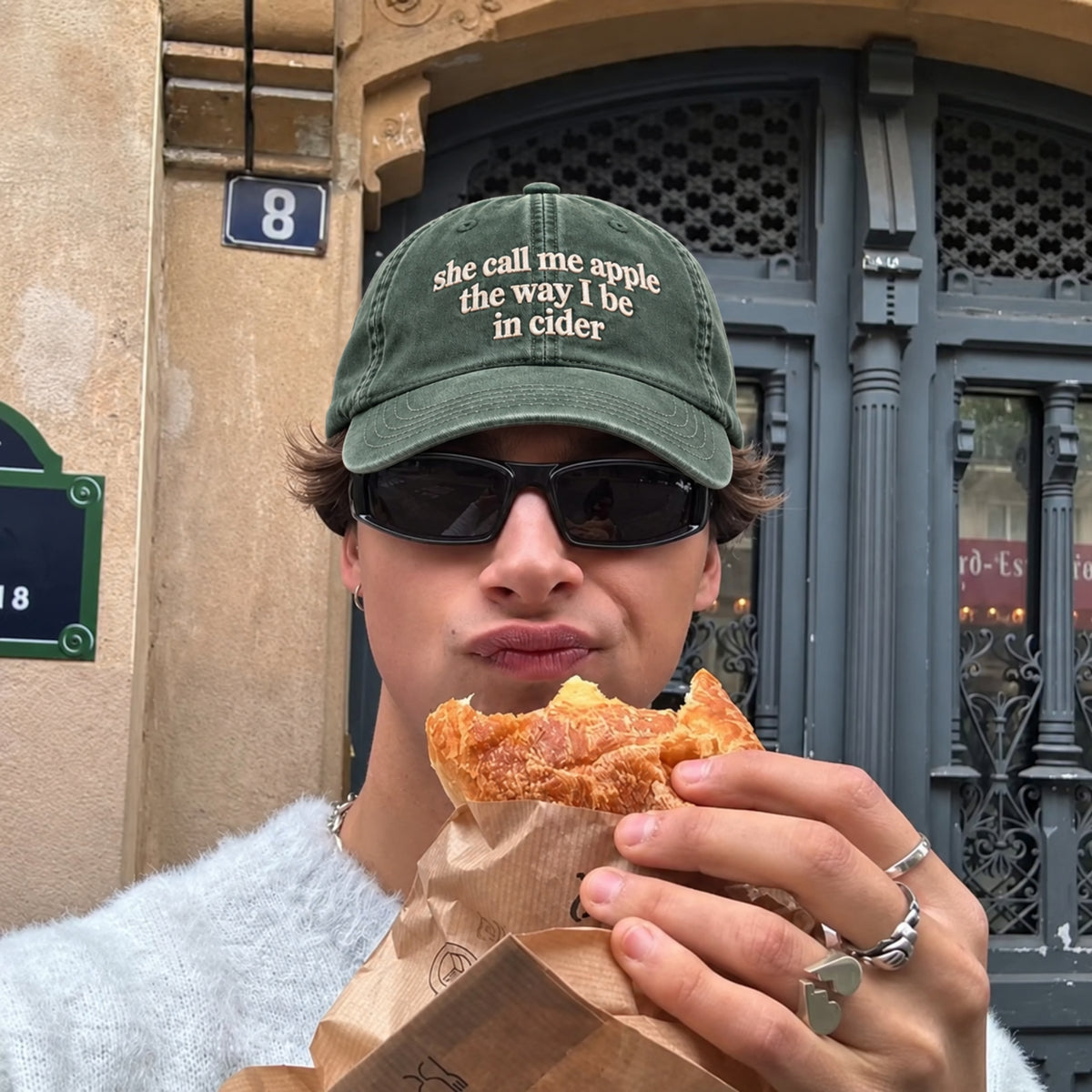 A person outdoors in front of a building entrance wearing a green baseball cap with the embroidered phrase 'she call me apple the way I be in cider' and holding a pastry in a brown paper bag.