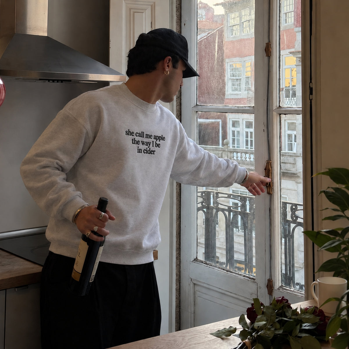 A man wearing a light gray sweatshirt with black text that reads 'she call me apple the way i be in elder' stands in a kitchen by a window, holding a wine bottle.
