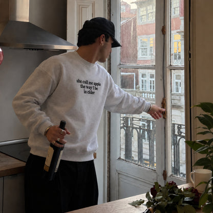 A man wearing a light gray sweatshirt with black text that reads 'she call me apple the way i be in elder' stands in a kitchen by a window, holding a wine bottle.