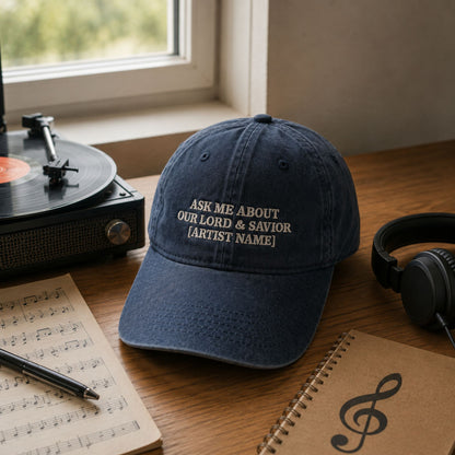 A navy blue baseball cap with white embroidered text reading 'ASK ME ABOUT OUR LORD & SAVIOR [ARTIST NAME]' sits on a wooden table in a music-themed setting, surrounded by a turntable, headphones, sheet music, and a notebook near a window.
