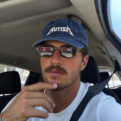 A man wearing a blue denim baseball cap with the word 'AUTISM' in white stylized lettering, sitting inside a car. He is also wearing dark sunglasses and a white t-shirt.