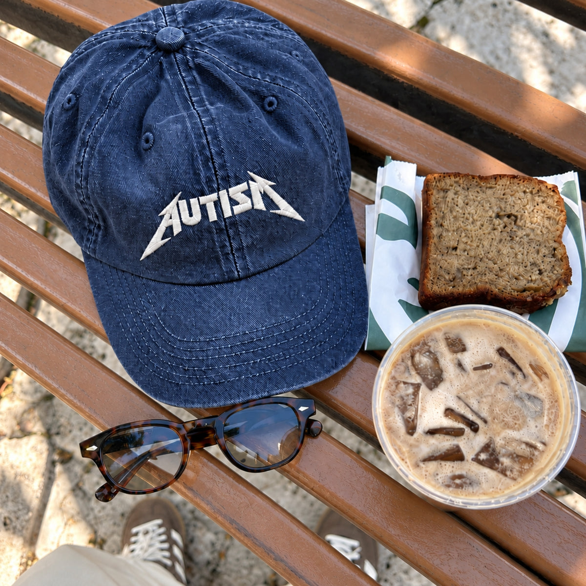 A navy blue baseball cap with a white 'AUTISM' logo on the front, styled on a wooden bench with a slice of banana bread, an iced coffee, sunglasses, and a shoe visible at the bottom.
