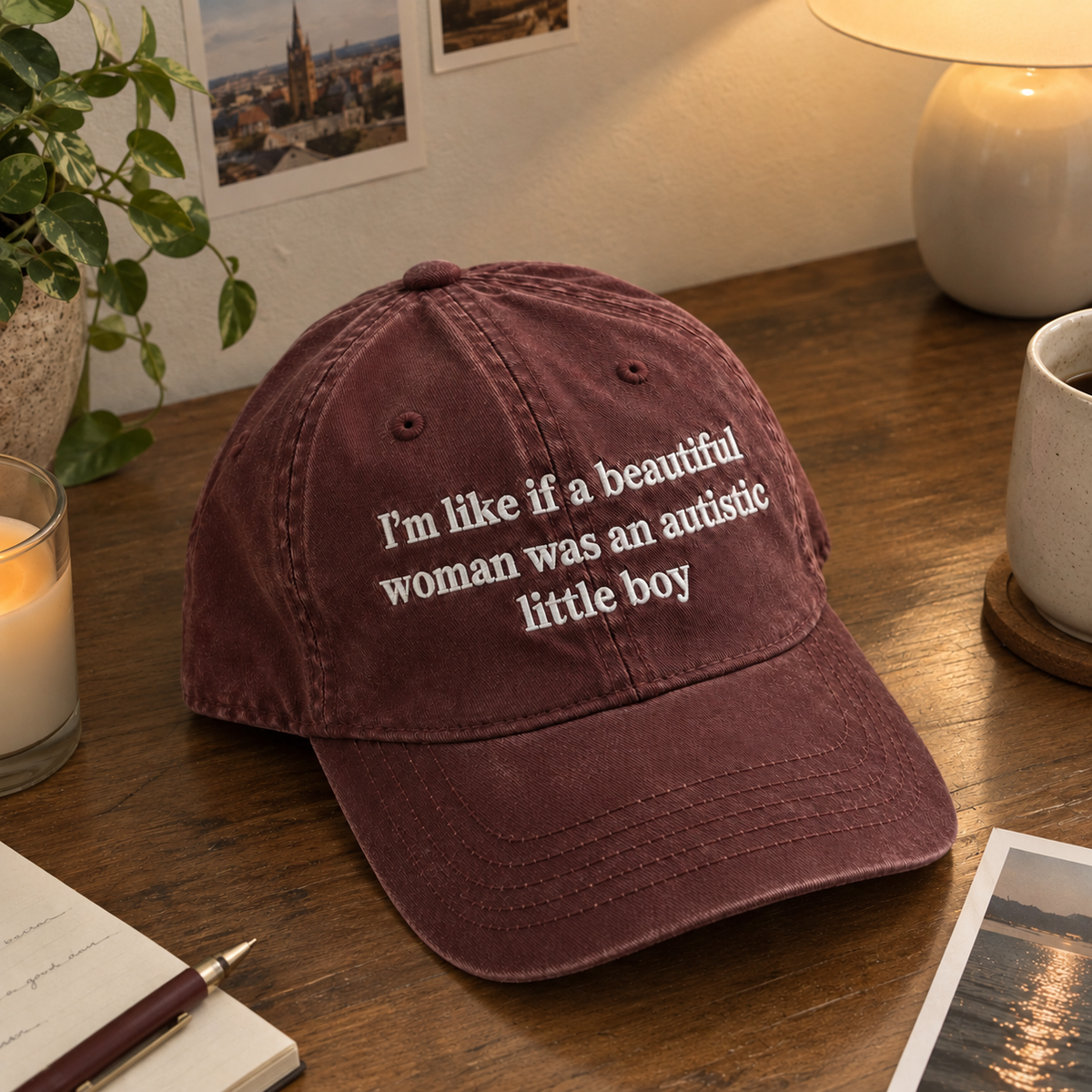 A maroon baseball cap with white embroidered text reading 'I'm like if a beautiful woman was an autistic little boy' sits on a wooden desk surrounded by cozy lifestyle props like a candle, mug, plant, and framed photos.