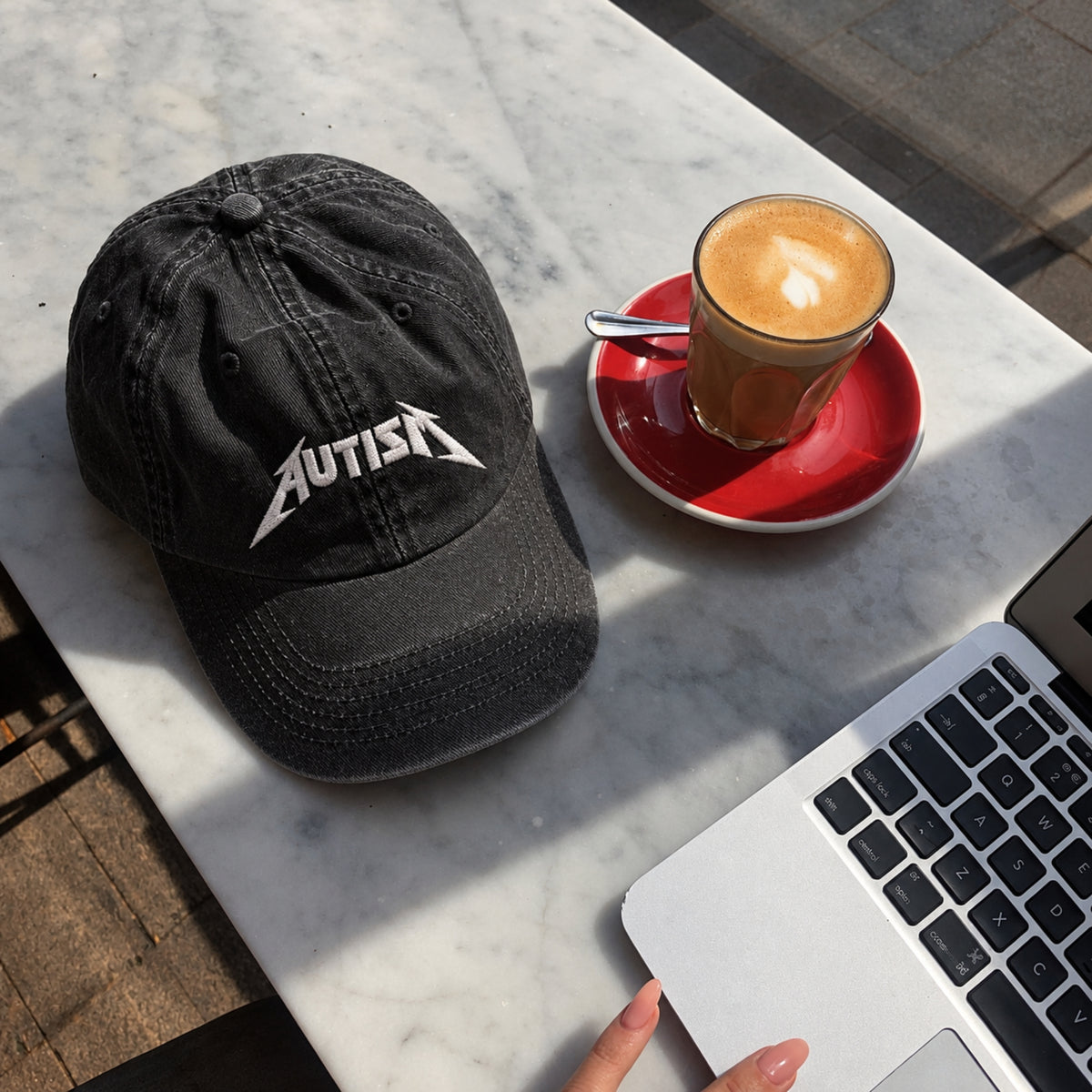 A black cotton baseball cap with a white embroidered 'AUTISM' logo in a stylized font, styled on a white marble table next to a cup of coffee and a laptop, with a hand visible at the bottom.