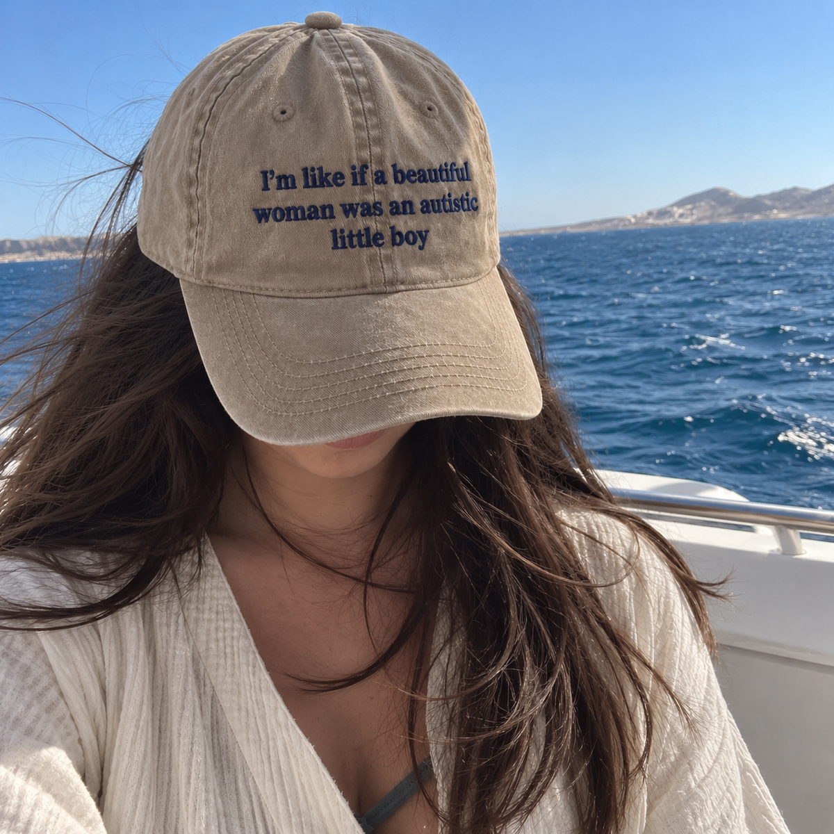 A woman on a boat wearing a beige baseball cap with dark blue embroidered text that reads, 'I'm like if a beautiful woman was an autistic little boy.'.