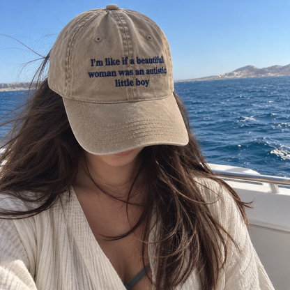 A woman on a boat wearing a beige baseball cap with dark blue embroidered text that reads, 'I'm like if a beautiful woman was an autistic little boy.'.