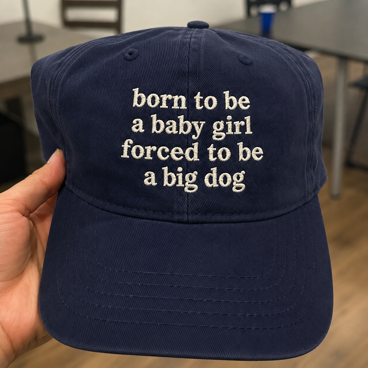 A navy blue baseball cap held by a hand, featuring white embroidered text that reads 'born to be a baby girl forced to be a big dog'.