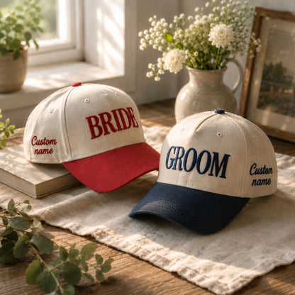 Two customizable baseball caps displayed on a rustic wooden table with a linen cloth, a book, and decorative flowers in the background. One cap is cream with a red brim and red embroidered text reading 'BRIDE Custom name', and the other is cream with a navy brim and navy embroidered text reading 'GROOM Custom name'.