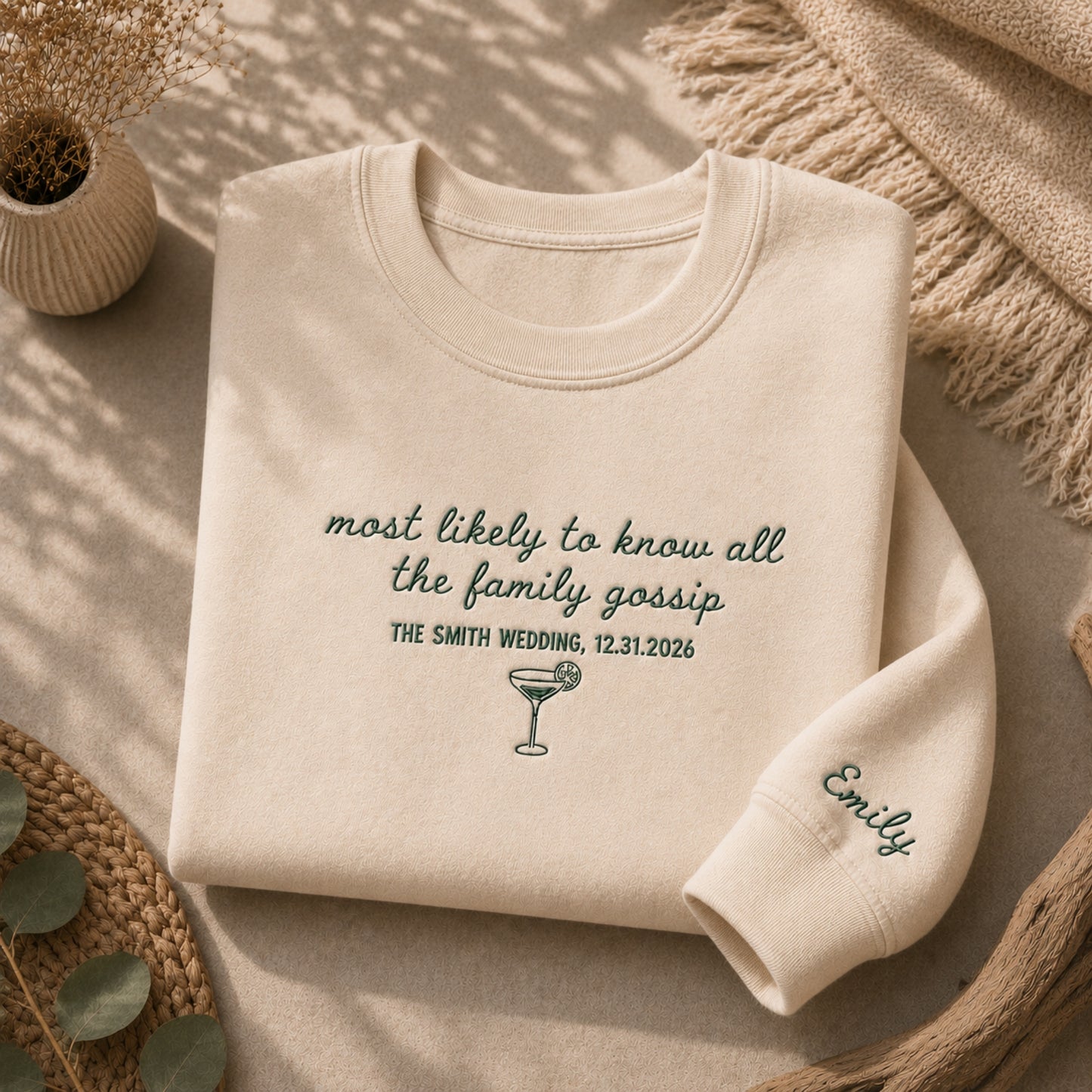 A folded cream sweatshirt with dark green embroidered text and a cocktail glass graphic, styled on a neutral surface with a knit blanket, woven mat, and small plant.