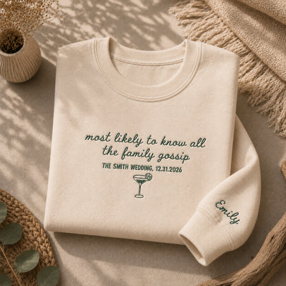 A folded cream sweatshirt with dark green embroidered text and a cocktail glass graphic, styled on a neutral surface with a knit blanket, woven mat, and small plant.