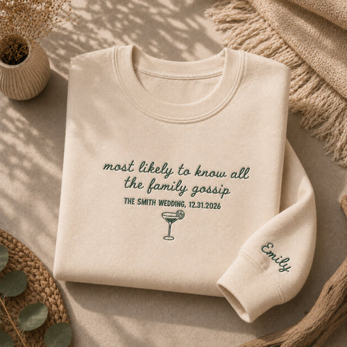 A folded cream sweatshirt with dark green embroidered text and a cocktail glass graphic, styled on a neutral surface with a knit blanket, woven mat, and small plant.