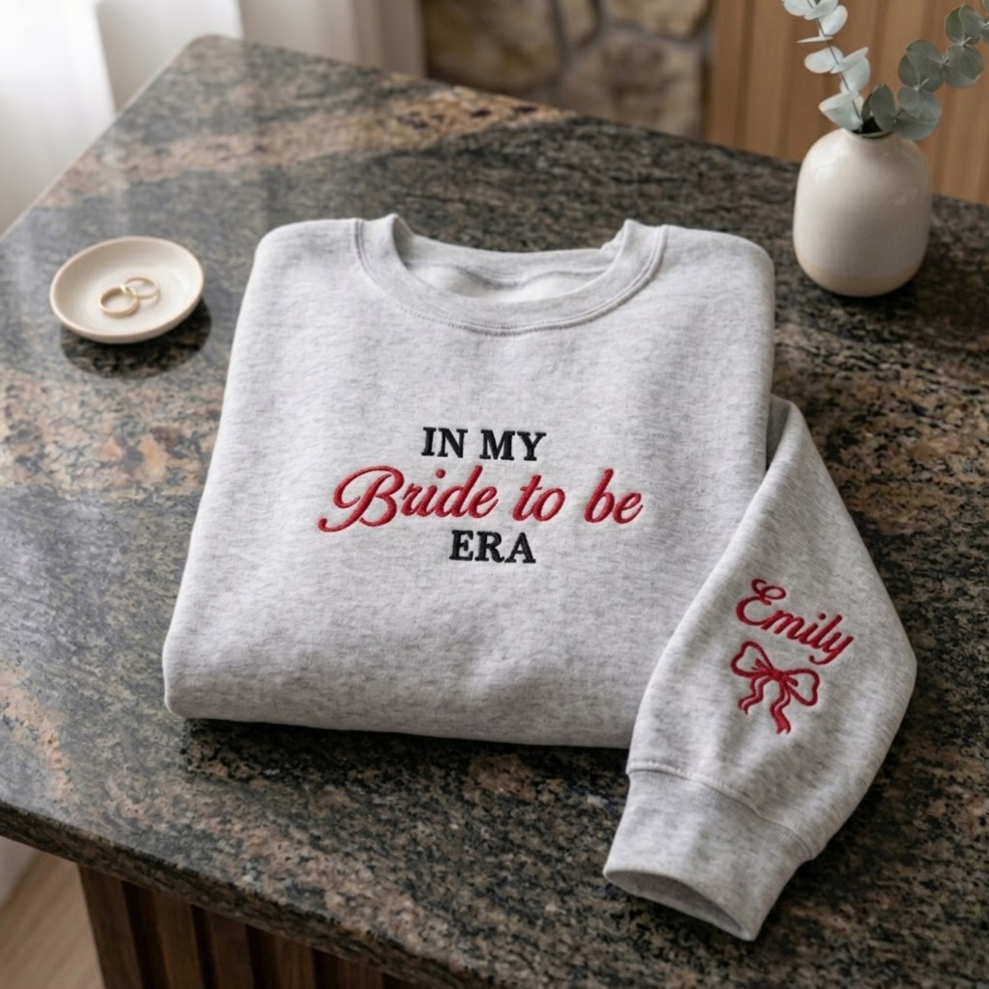 A folded light heather gray sweatshirt on a granite countertop, printed with the phrase 'IN MY Bride to be ERA' in black and red lettering. The right sleeve has 'Emily' and a red bow printed on it.