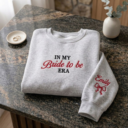 A folded light heather gray sweatshirt on a granite countertop, printed with the phrase 'IN MY Bride to be ERA' in black and red lettering. The right sleeve has 'Emily' and a red bow printed on it.