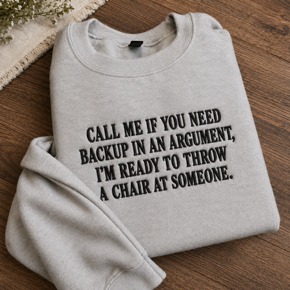 A folded light heather gray sweatshirt with bold black text reading 'CALL ME IF YOU NEED BACKUP IN AN ARGUMENT, I'M READY TO THROW A CHAIR AT SOMEONE.' displayed on a wooden surface with a small decorative textile corner visible.