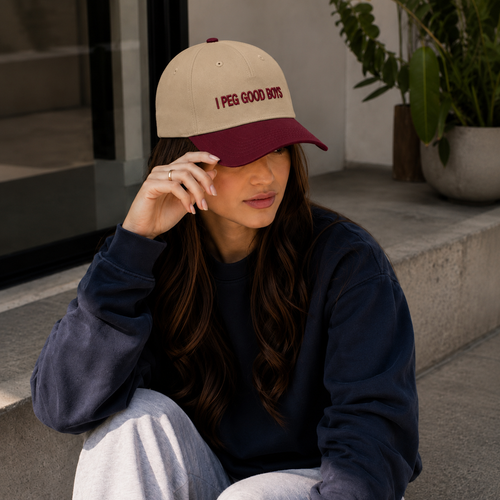 A young woman sits outdoors on concrete steps, wearing a beige-and-burgundy baseball cap with the words “I PEG GOOD BOYS,” a dark navy sweatshirt, and light gray pants. She has long dark hair and rests one hand near the brim of her cap. Plants and a modern building entrance are visible in the background.