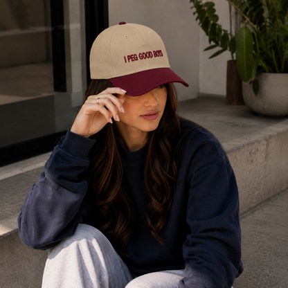 A young woman sits outdoors on concrete steps, wearing a beige-and-burgundy baseball cap with the words “I PEG GOOD BOYS,” a dark navy sweatshirt, and light gray pants. She has long dark hair and rests one hand near the brim of her cap. Plants and a modern building entrance are visible in the background.