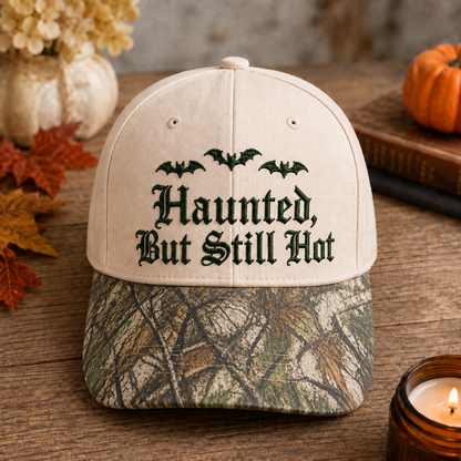 A baseball cap with a cream-colored crown and a green/brown camouflage brim, featuring the text 'Haunted, But Still Hot' in black gothic font with three bats above it. The cap is styled on a rustic wooden table with autumn leaves, a candle, a book, and a small pumpkin in the background.