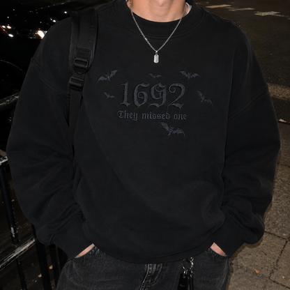A person wearing a black crewneck sweatshirt with a dark gray gothic graphic that reads '1692 They missed one' surrounded by small bat silhouettes. The person is also wearing a silver chain necklace and a black backpack strap, standing outdoors at night.