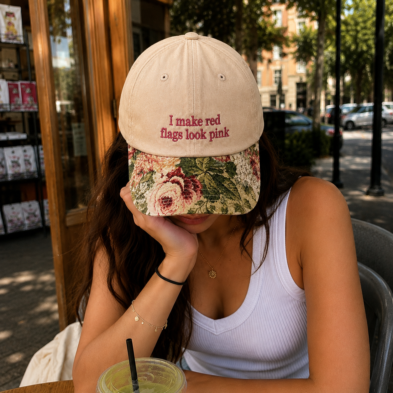 Woman wearing a beige embroidered baseball hat with pink text 'I make red flags look pink' and a floral brim, sitting at an outdoor cafe.