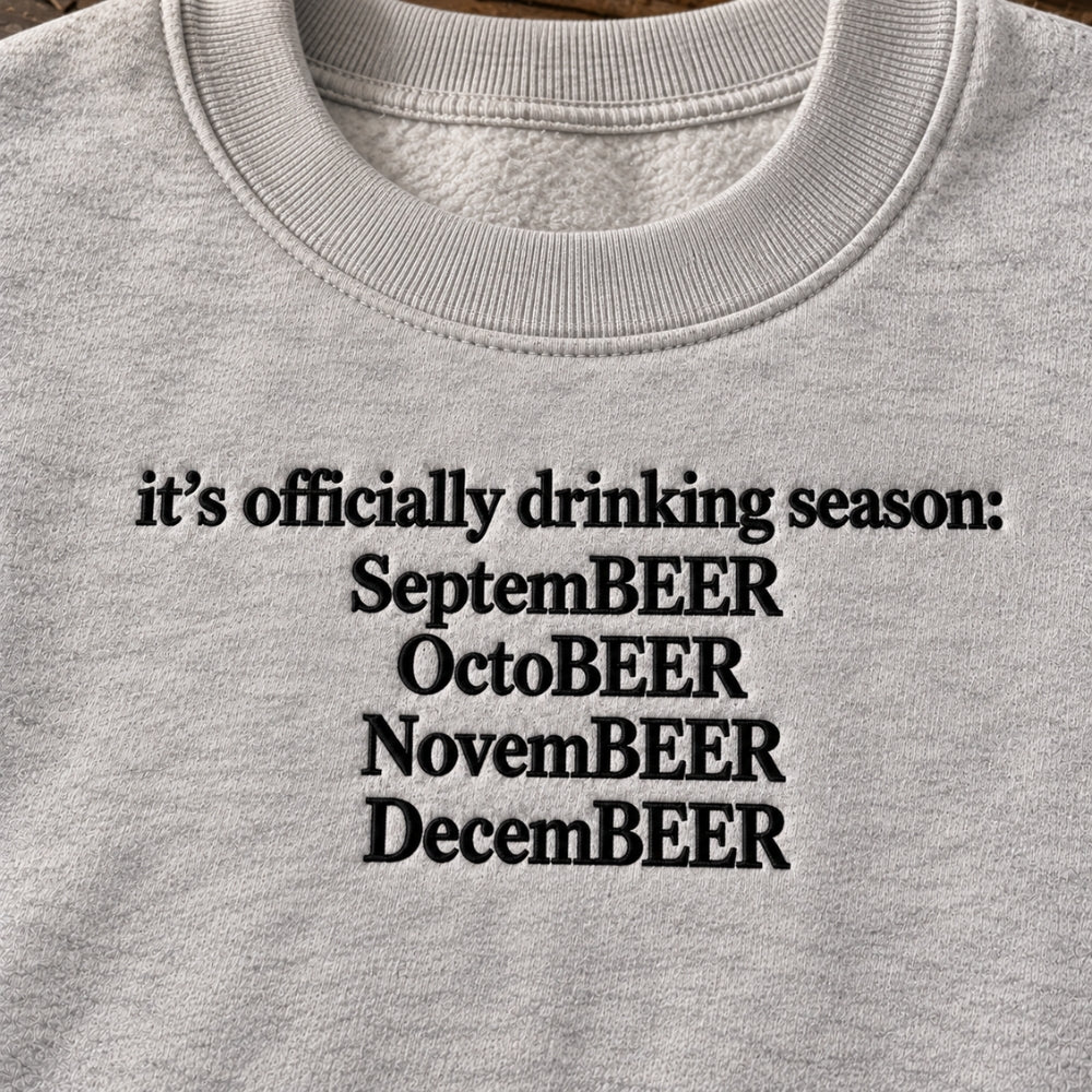 Close-up of a light heather gray crewneck sweatshirt featuring black embroidered text that reads 'it's officially drinking season: SeptemBEER OctoBEER NovemBEER DecemBEER'.