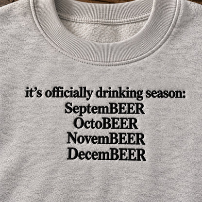 Close-up of a light heather gray crewneck sweatshirt featuring black embroidered text that reads 'it's officially drinking season: SeptemBEER OctoBEER NovemBEER DecemBEER'.