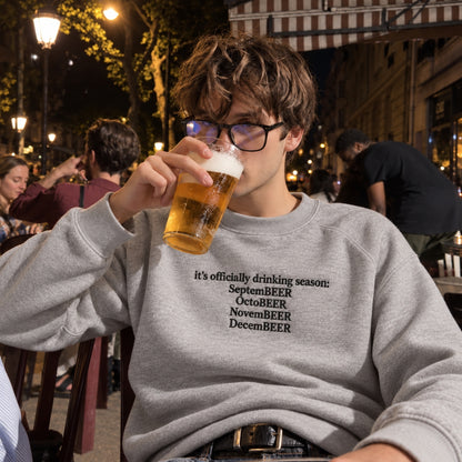 A man wearing a light gray sweatshirt with black text reading 'it's officially drinking season: SeptemBEER OctoBEER NovemBEER DecemBEER' sits at an outdoor bar or cafe at night, holding a glass of beer.
