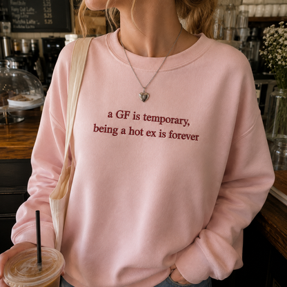Woman standing indoors holding an iced drink, wearing a light pink crewneck sweatshirt with dark pink embroidered text that reads "a GF is temporary, being a hot ex is forever".