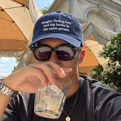 A man wearing a navy blue baseball cap with the white embroidered text 'imagine finding love and big boobs in the same person' and black sunglasses, drinking from a glass outdoors under beige patio umbrellas.