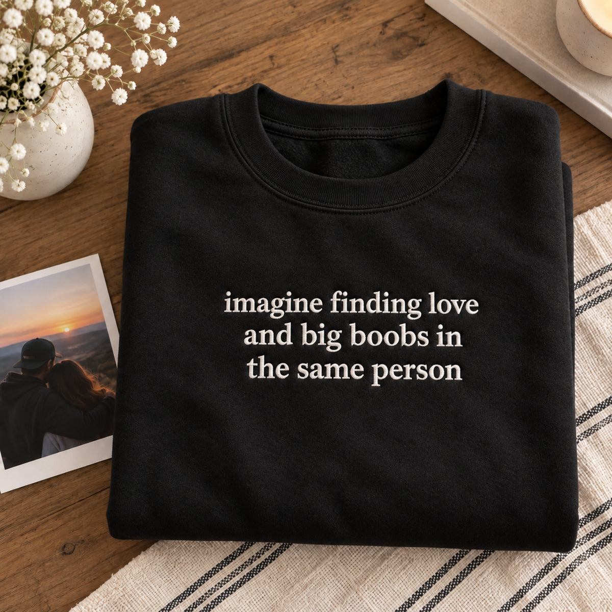 alt text
Folded black embroidered sweatshirt featuring the quote “imagine finding love and big boobs in the same person,” styled on a wooden table with flowers and a couple photo.