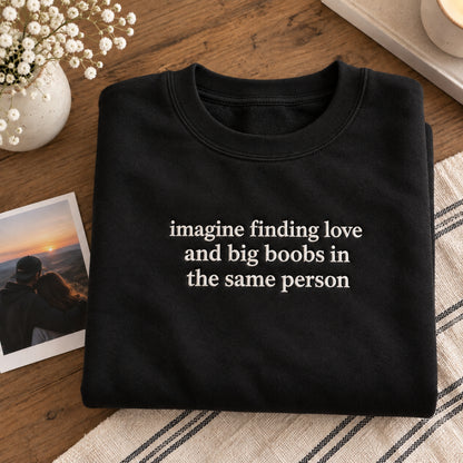 alt text
Folded black embroidered sweatshirt featuring the quote “imagine finding love and big boobs in the same person,” styled on a wooden table with flowers and a couple photo.