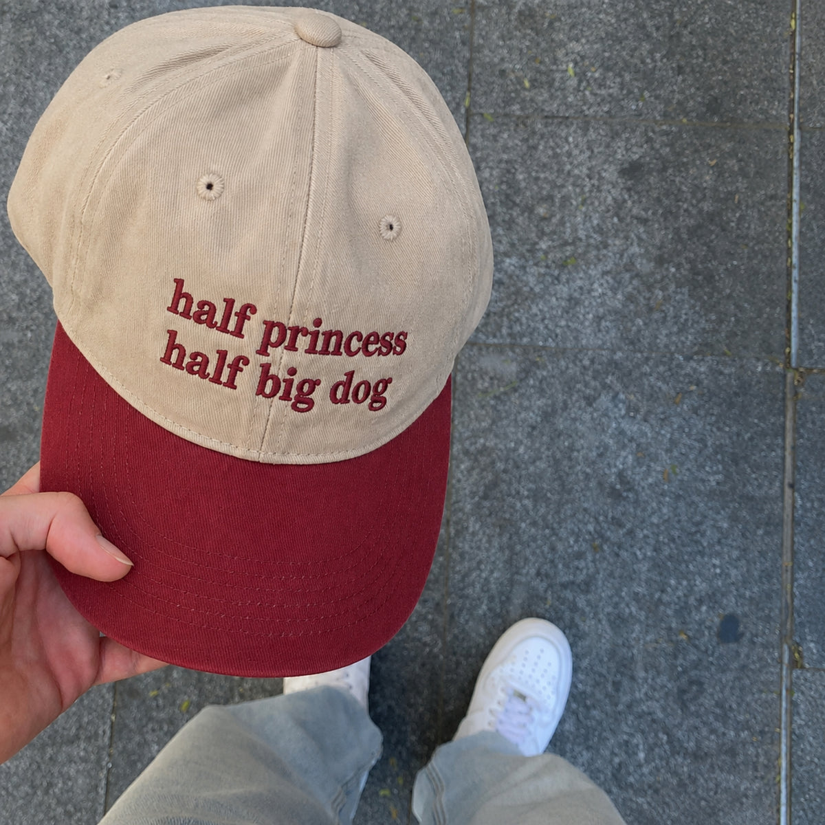 A person holds a beige and burgundy baseball cap with the embroidered text 'half princess half big dog' in burgundy. The background shows gray pavement and the person's white sneakers and jeans.