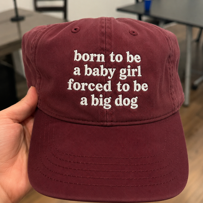 A hand holding a maroon baseball cap with white embroidered text that reads 'born to be a baby girl forced to be a big dog' in a real indoor setting.