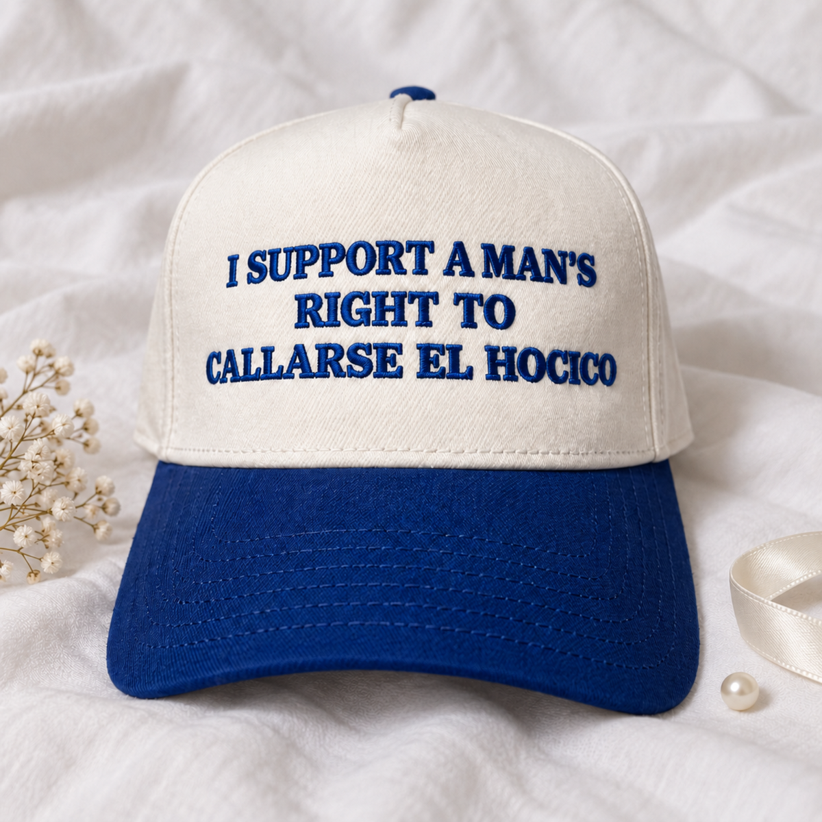A baseball cap with an off-white crown and a royal blue brim, featuring the Spanish text 'I SUPPORT A MAN'S RIGHT TO CALLARSE EL HOCICO' embroidered in blue on the front. The cap is styled on a white fabric background with baby's breath and a pearl.