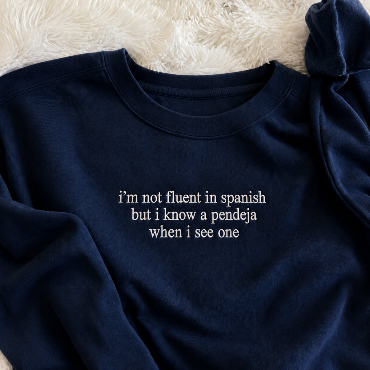 A navy blue crewneck sweatshirt with white embroidered text reading 'i'm not fluent in spanish but i know a pendeja when i see one', laid flat on a white faux fur surface.