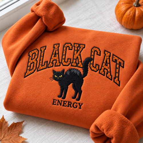 An orange fleece sweatshirt is folded and draped on a light fabric surface, featuring a black embroidered cat with yellow eyes and the words 'BLACK CAT ENERGY' in black outlined lettering. A small pumpkin, autumn leaf, and window sill are visible around it.