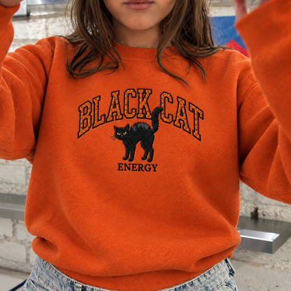 A person wearing a bright orange sweatshirt with a black cat graphic and the words 'BLACK CAT ENERGY' on the front, paired with light blue denim jeans.