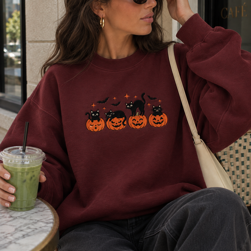 A woman wearing a maroon crewneck sweatshirt with a Halloween graphic of black cats on jack-o'-lanterns, sitting at a cafe and holding a green iced coffee.