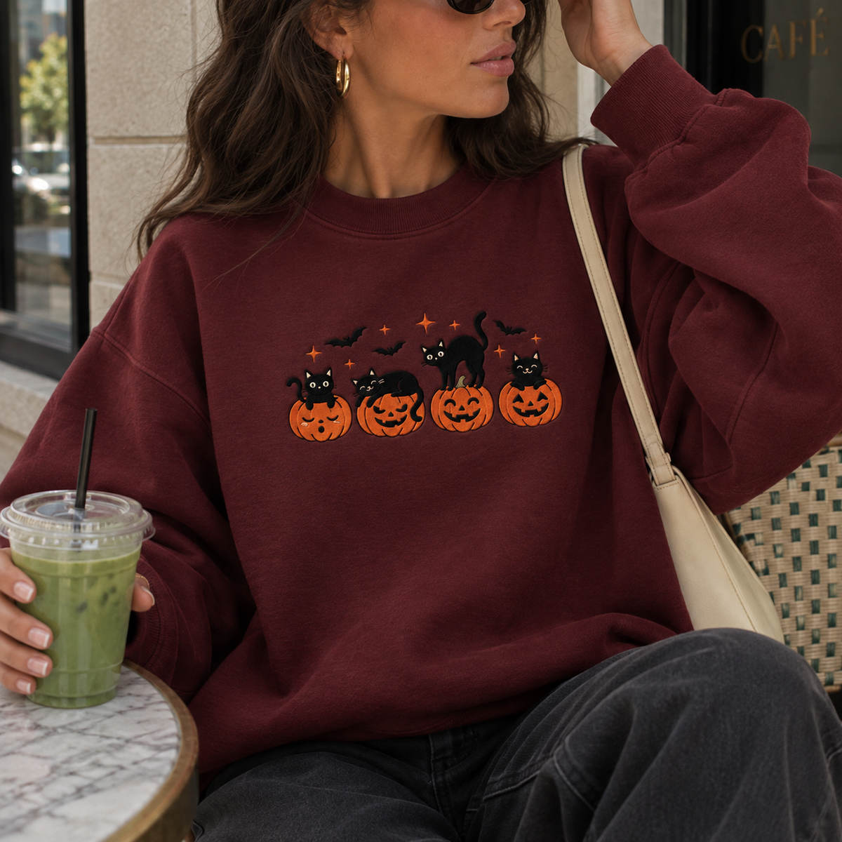 A woman wearing a maroon crewneck sweatshirt with a Halloween graphic of black cats on jack-o'-lanterns, sitting at a cafe and holding a green iced coffee.