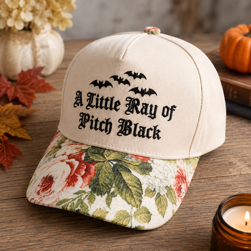 A cream-colored baseball cap with 'A Little Ray of Pitch Black' embroidered in black gothic font under five black bats. The brim features a vintage-style floral print with red roses and green leaves on a cream background.