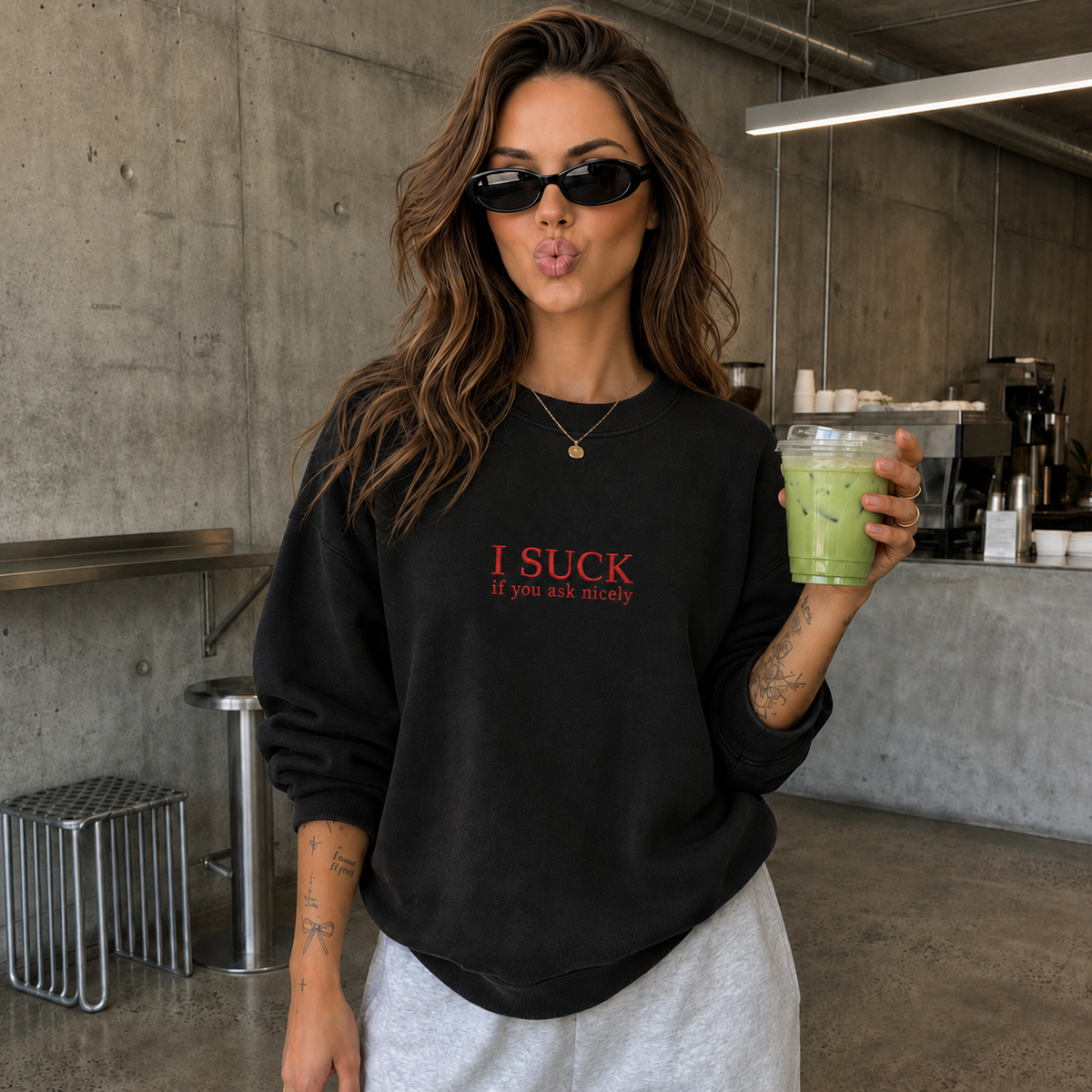 A woman wearing a black crewneck sweatshirt with red text that reads 'I SUCK If you ask nicely' and light gray sweatpants, holding a green iced drink in a modern concrete-walled cafe setting.