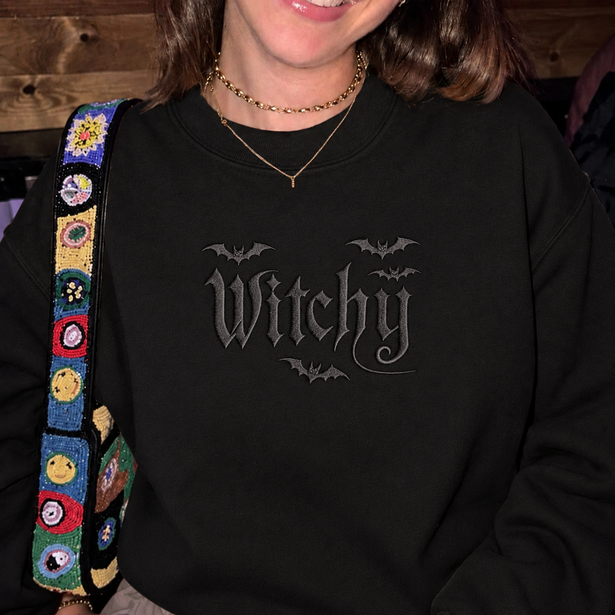 A person wearing a black crewneck sweatshirt with a gray gothic-style 'Witchy' graphic and bat motifs on the chest. The person is also wearing gold necklaces and has a colorful beaded shoulder strap visible on the left side.